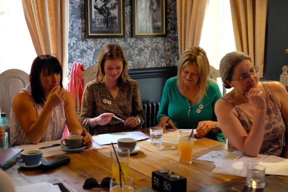 A group of four women engaged in discussion around a table with drinks and papers.