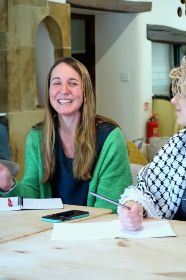 Woman with long hair in a green cardigan smiles while sitting at a table during a discussion.