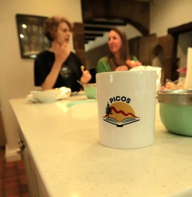 Two women chatting at a kitchen counter with bowls and a mug in the foreground.
