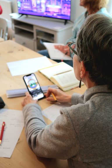 A person using a smartphone at a table with papers and a TV in the background.